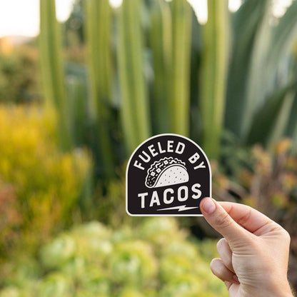 A hand holds the Fueled By Tacos sticker in front of tall cactus and succulents at golden hour.