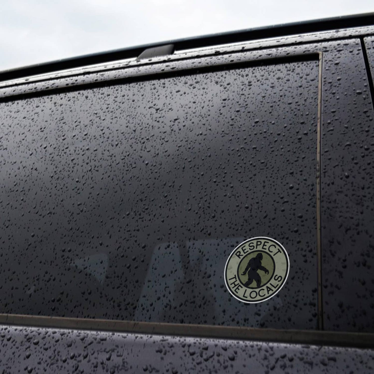 Rain beads across a car window where the Bigfoot Respect The Locals sticker clings near the door frame.