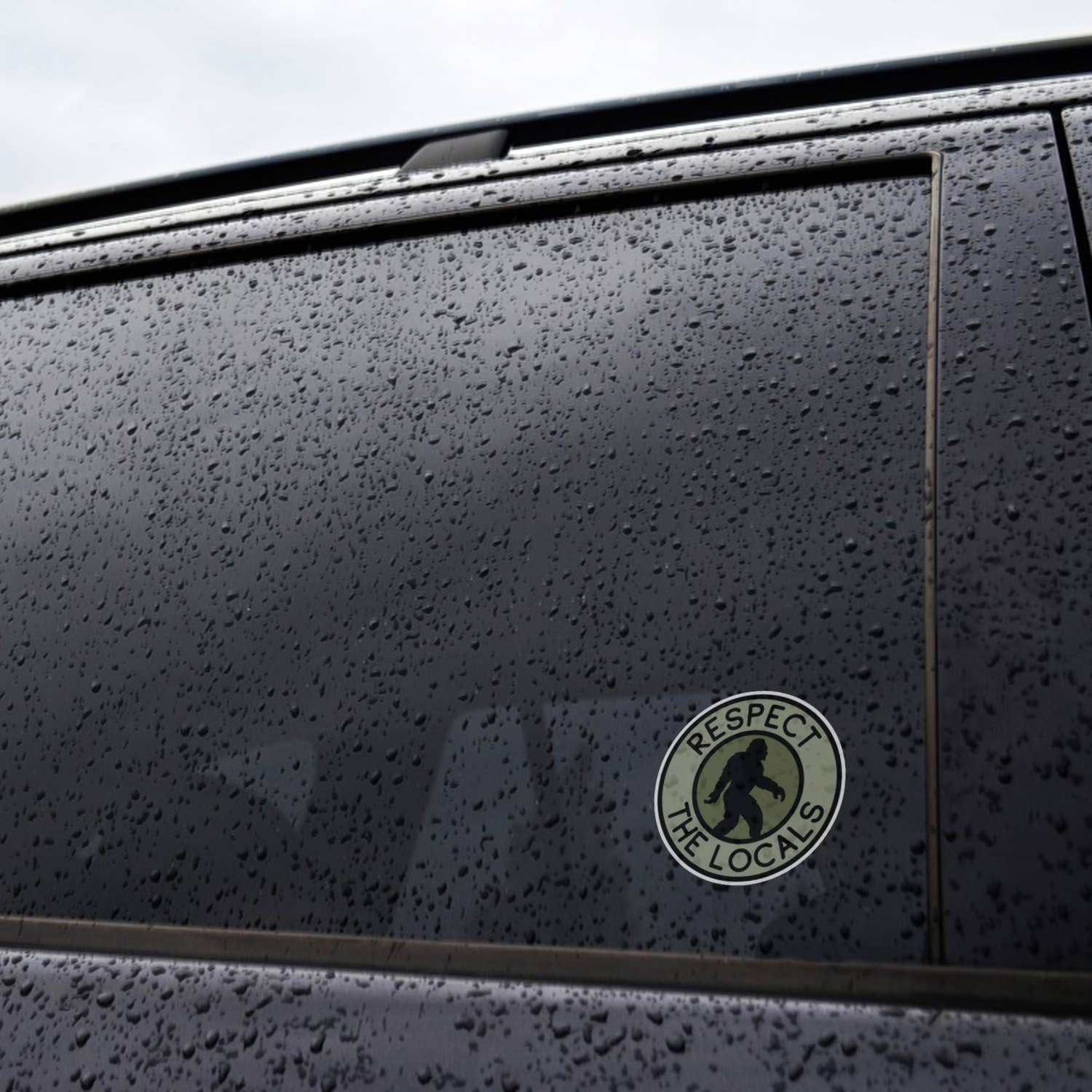 Rain beads across a car window where the Bigfoot Respect The Locals sticker clings near the door frame.