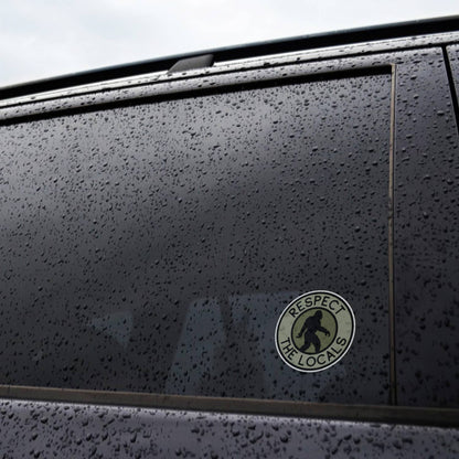 Rain beads across a car window where the Bigfoot Respect The Locals sticker clings near the door frame.
