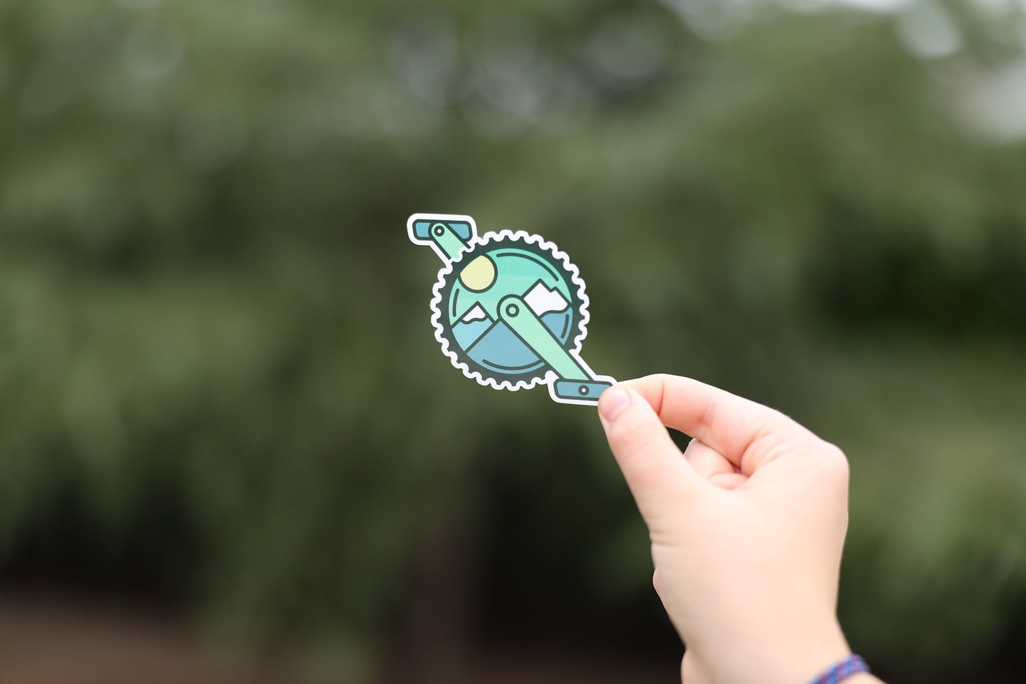 Held at arm's length against dark trees, the Mountain Bike Crank sticker shows the full chainring and crank arms.