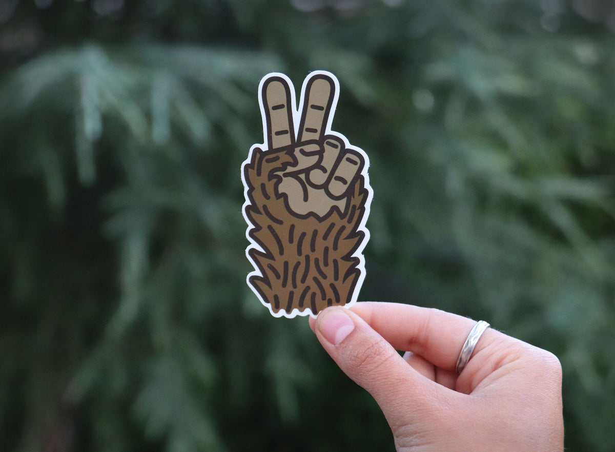 Fingers with a silver ring hold a furry Sasquatch peace sign sticker against pine needles.
