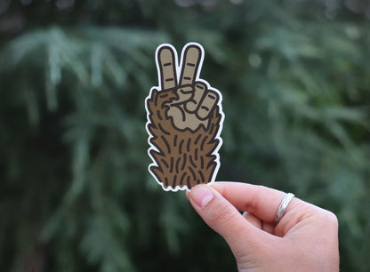 Fingers with a silver ring hold a furry Sasquatch peace sign sticker against pine needles.