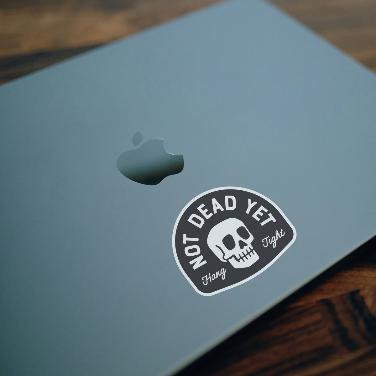 On the grey lid of a laptop, the Not Dead Yet sticker sits near the indented Apple logo.