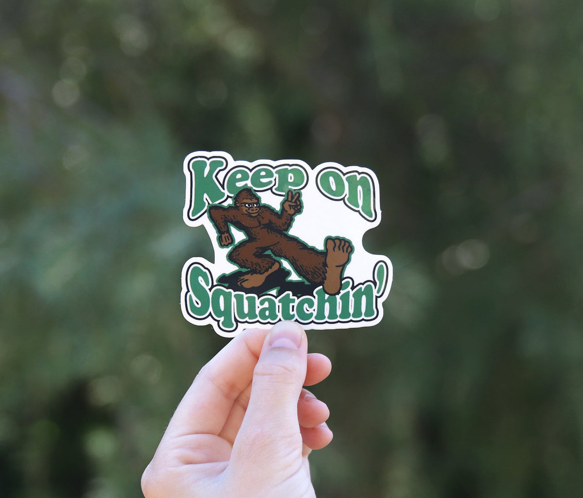 Backed by blurred green branches, the Keep on Squatchin sticker sits smaller in frame between two fingers.