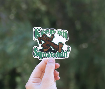 Backed by blurred green branches, the Keep on Squatchin sticker sits smaller in frame between two fingers.
