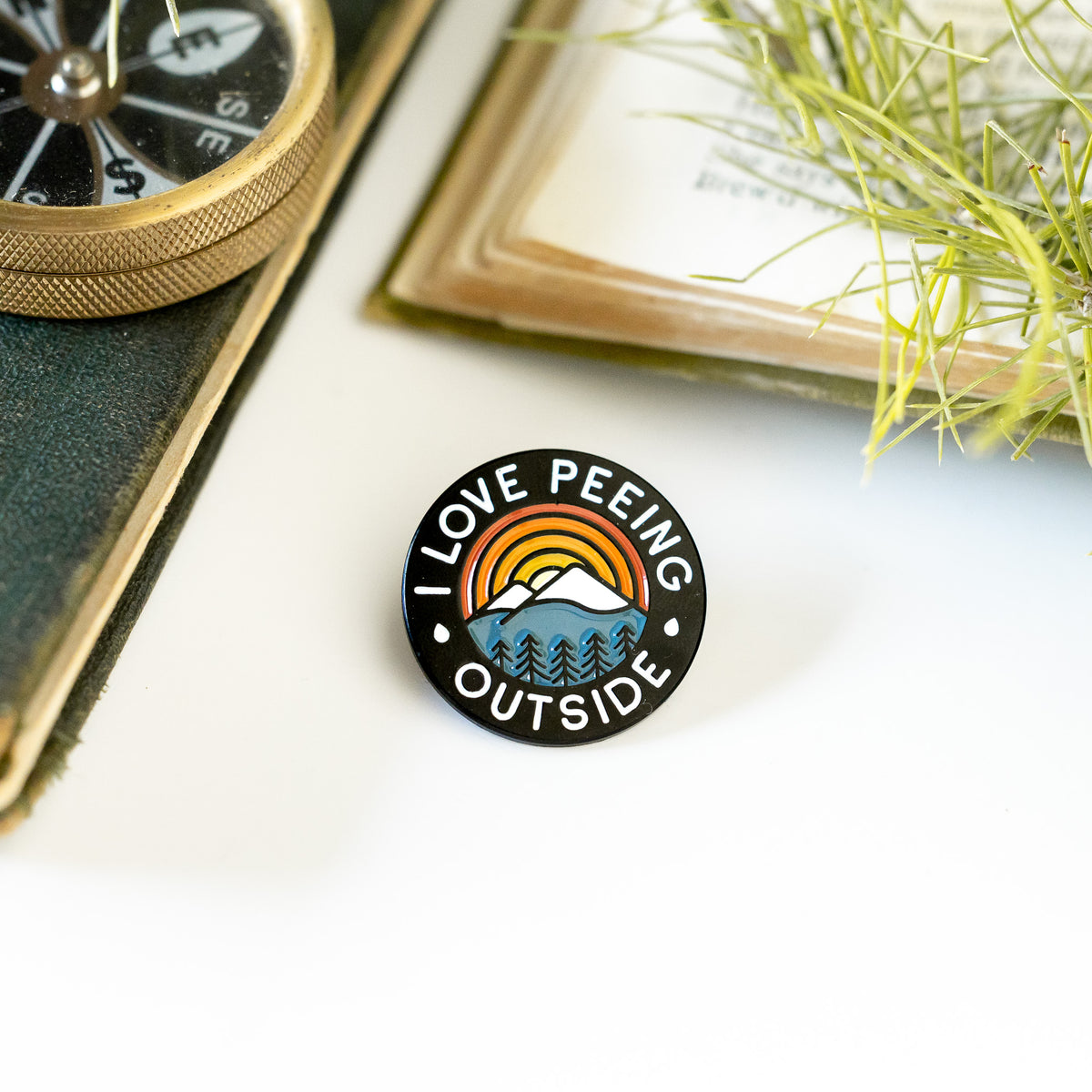 Beside an open compass and a worn journal, the I Love Peeing Outside pin rests on a white surface near dried grass.