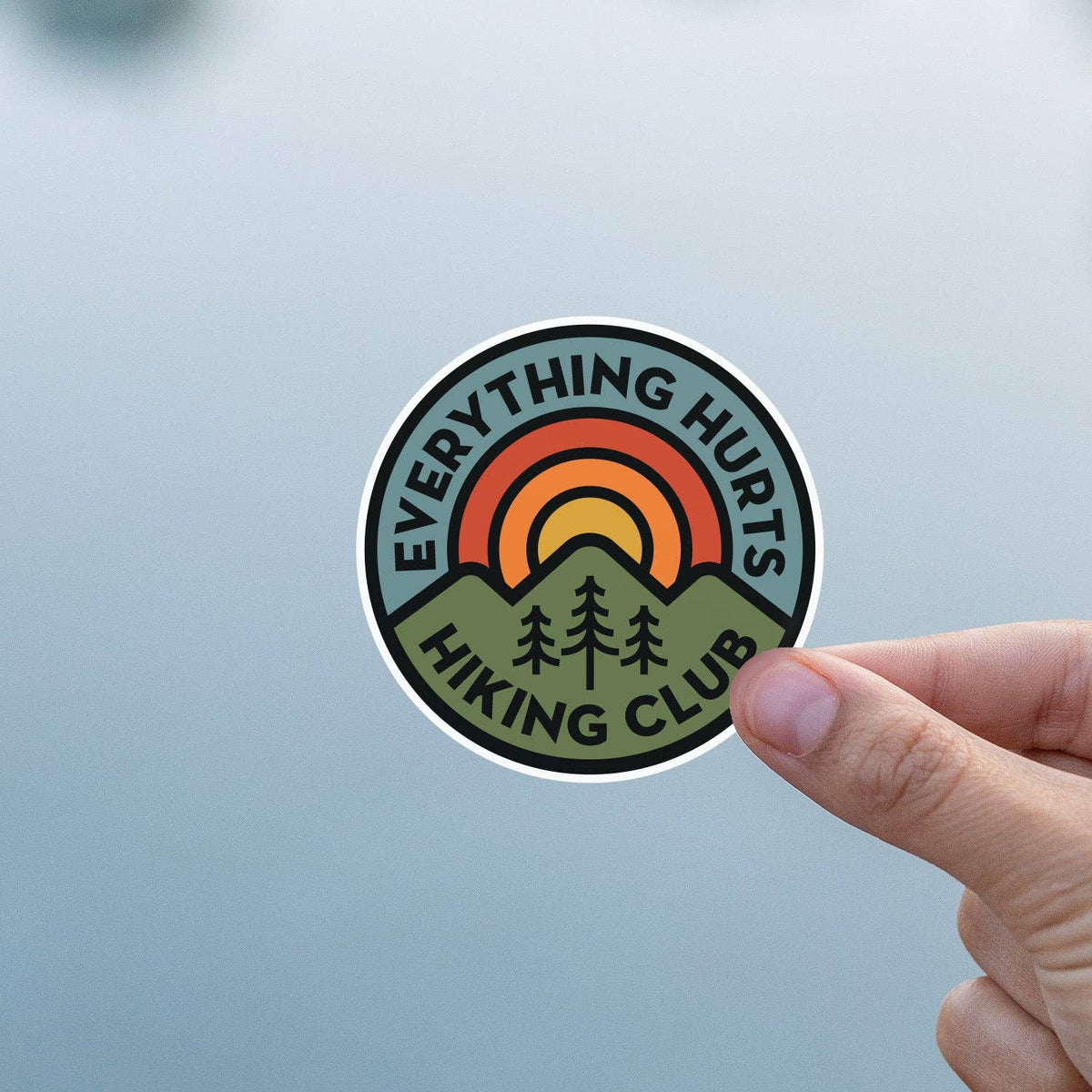 Against a plain pale blue background, fingers hold the Everything Hurts Hiking Club sticker upright.