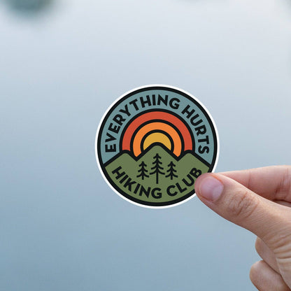 Against a plain pale blue background, fingers hold the Everything Hurts Hiking Club sticker upright.