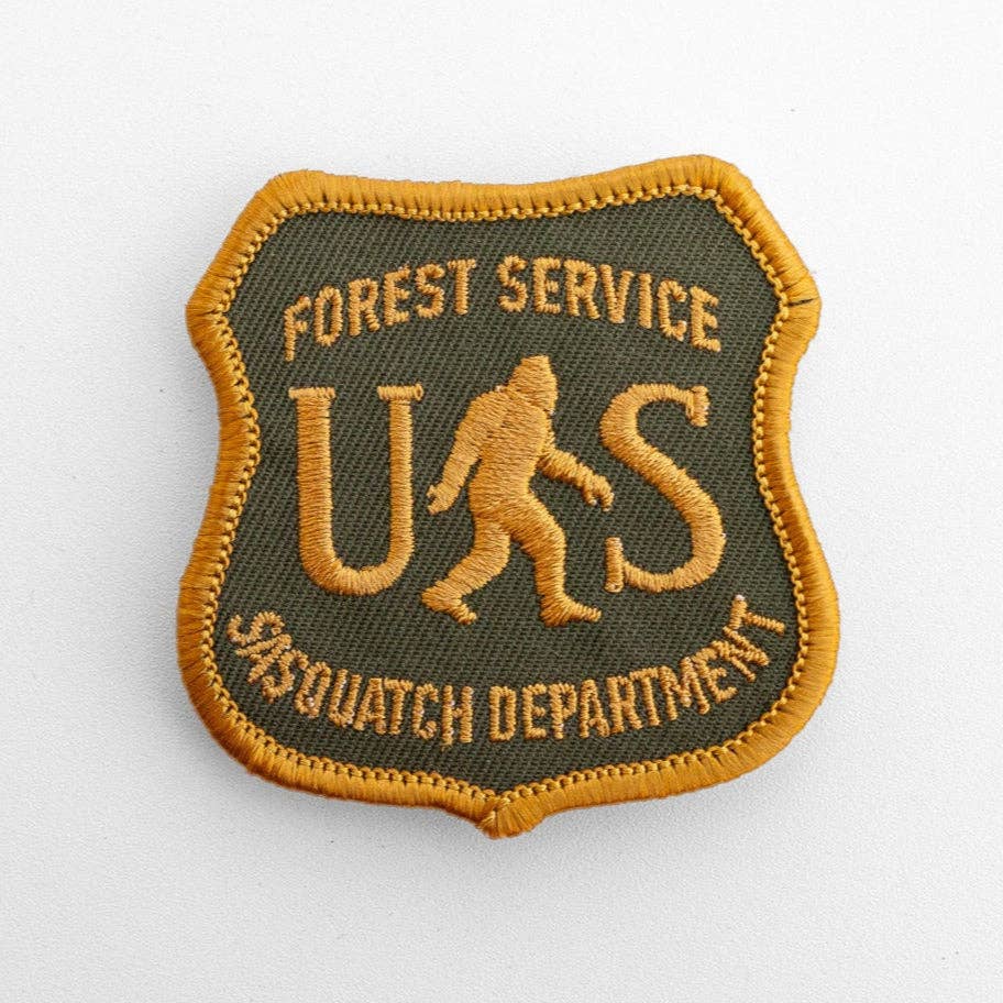 Flat on white, the US Sasquatch Department patch shows gold shield stitching over olive twill with a walking bigfoot.