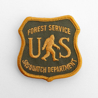 Flat on white, the US Sasquatch Department patch shows gold shield stitching over olive twill with a walking bigfoot.