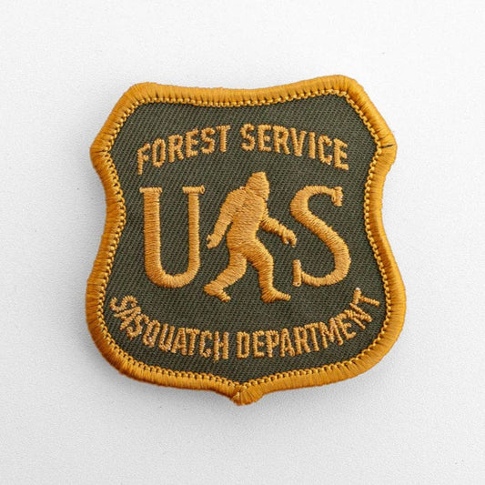 Flat on white, the US Sasquatch Department patch shows gold shield stitching over olive twill with a walking bigfoot.