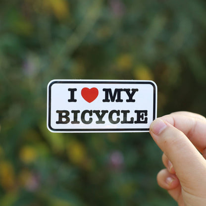 Fingers hold the I Heart My Bicycle sticker with its red heart against blurred green and yellow foliage.
