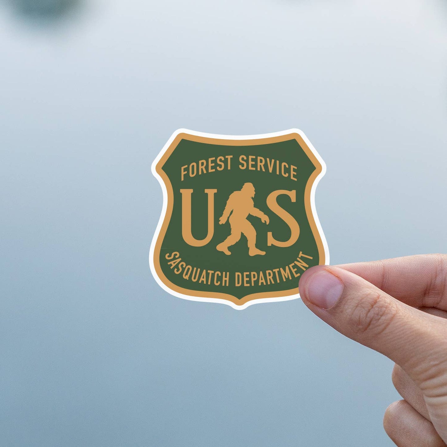 Against a soft grey blue background, a thumb steadies the US Sasquatch Department sticker.