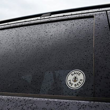 Rain beads across a dark car window beside the Save The Bees sticker applied near the frame.