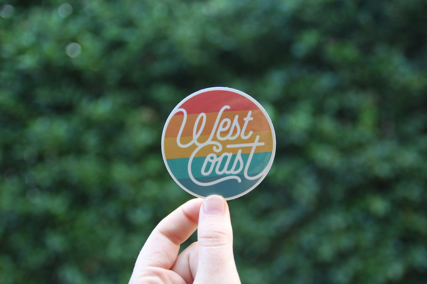 Held a little farther back against dark leaves, the West Coast Life sticker catches soft daylight.