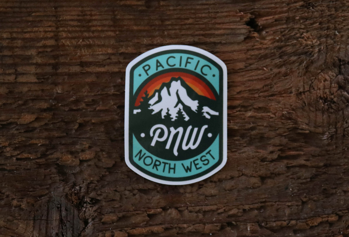 Stuck flat on rough brown wood, the PNW Compass sticker shows its mountain, rainbow arc and teal border.