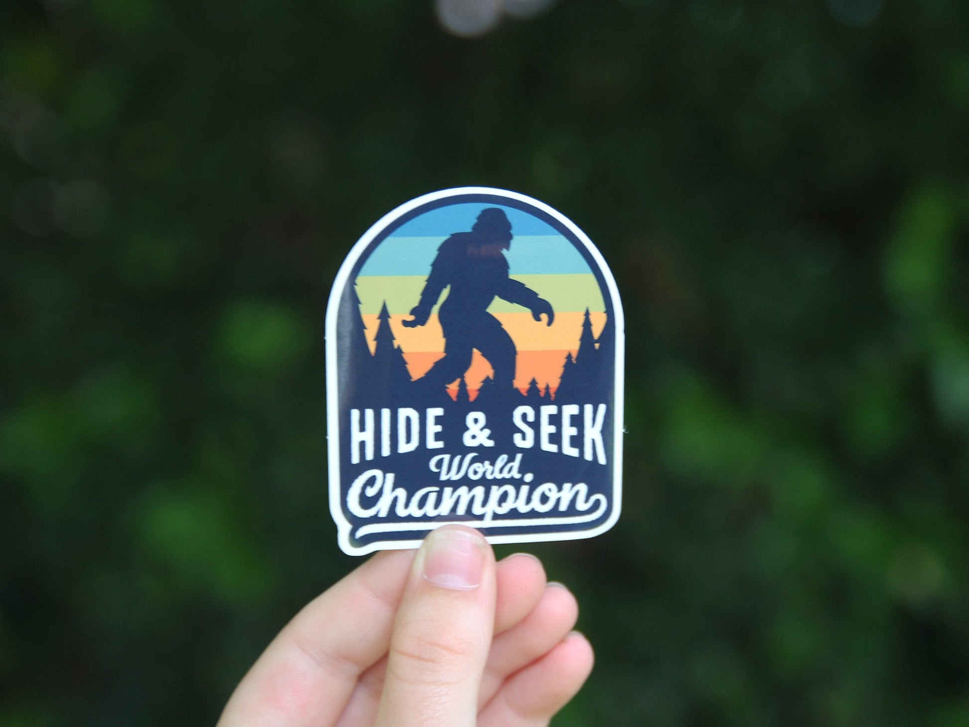 Fingertips lift the Bigfoot Hide & Seek World Champion sticker in front of a shadowed green hedge.