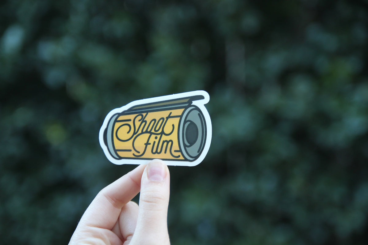 Held a little lower against the same dark shrubs, the Shoot Film sticker shows its lens and script lettering.
