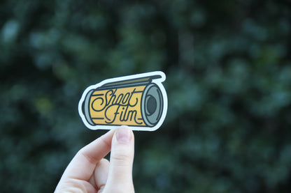 Held a little lower against the same dark shrubs, the Shoot Film sticker shows its lens and script lettering.