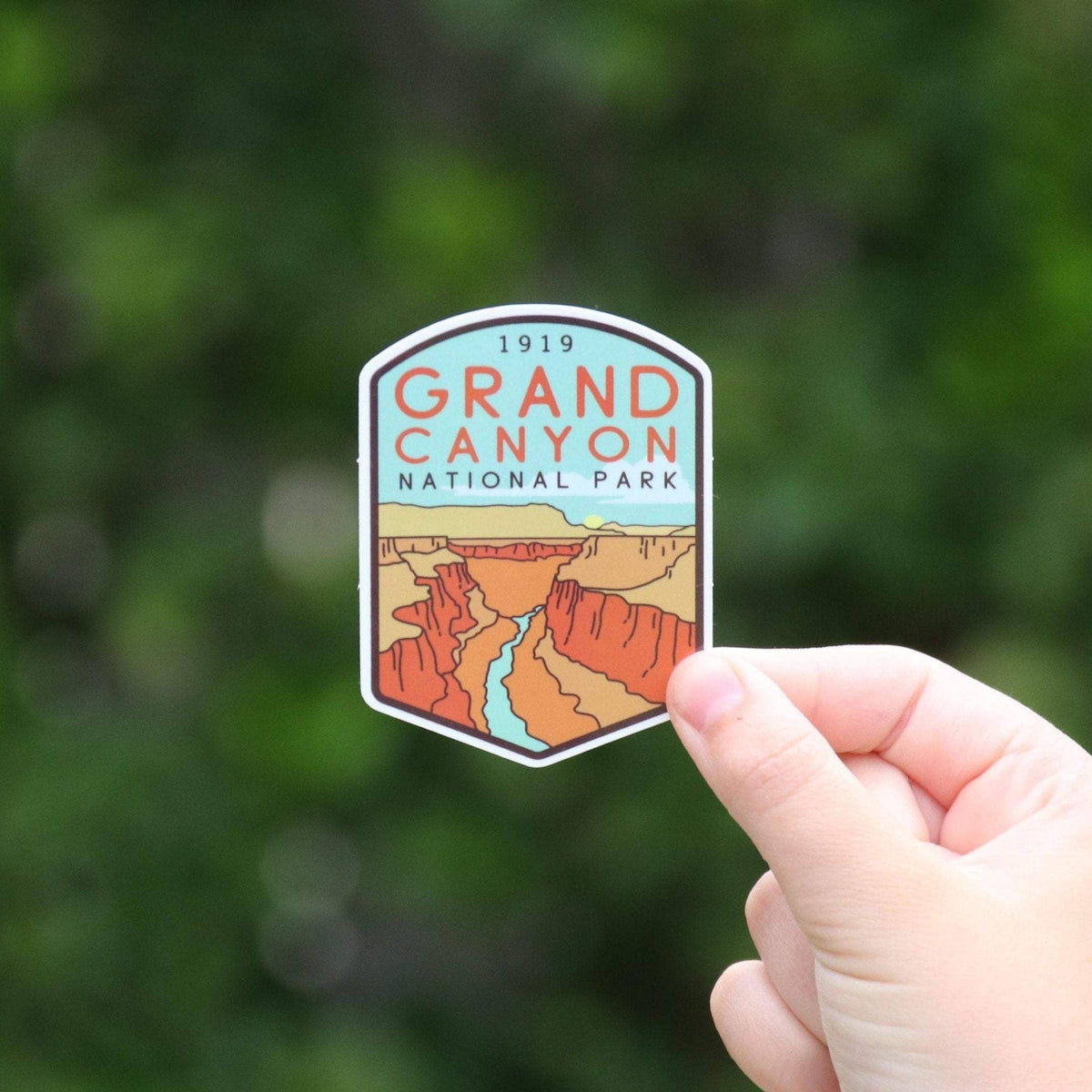 A hand holds the Grand Canyon National Park sticker against blurred green leaves, canyon walls and river showing.