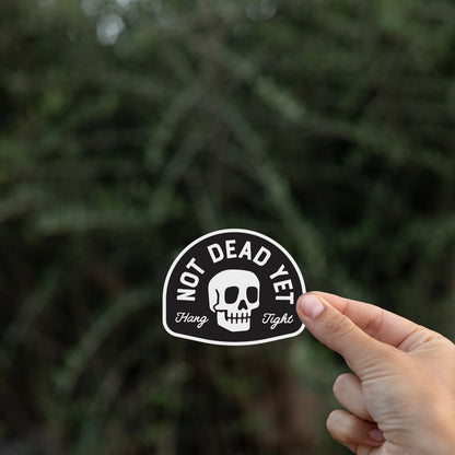 Against blurred dark branches, a hand raises the Not Dead Yet sticker by its lower edge.