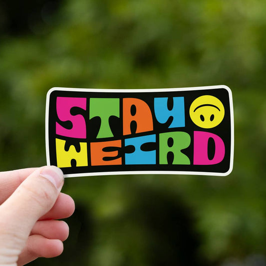 A hand holds up the Stay Weird sticker, its bright block letters and smiley face bold against blurred green leaves.