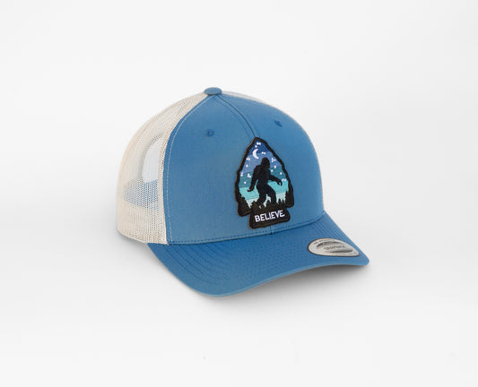 Angled on white, the blue and cream Bigfoot Believe trucker hat shows its arrowhead patch and starry night scene.