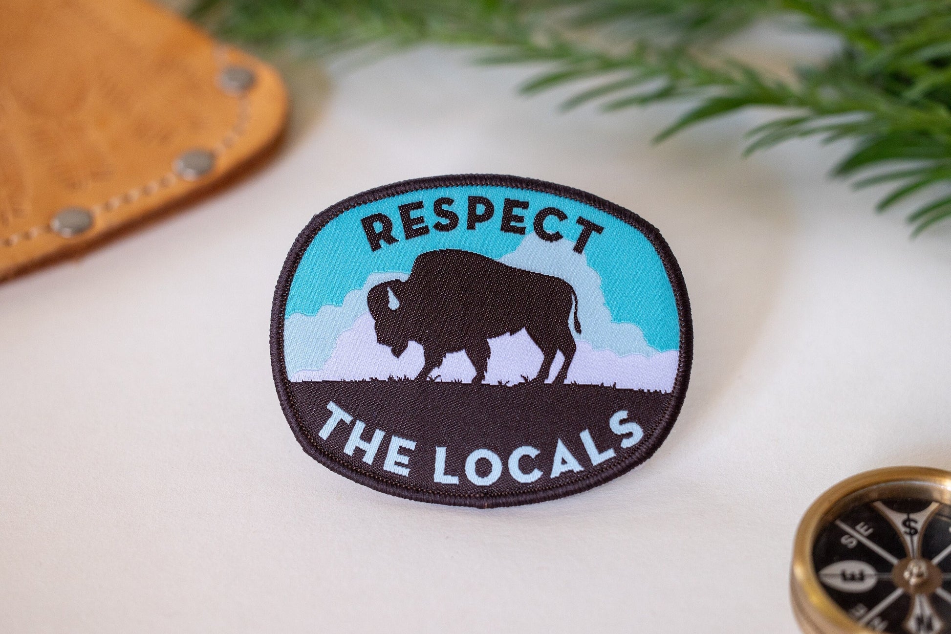Laid on cream fabric near a brass compass and leather pouch, the Respect The Locals bison patch shows its teal sky.