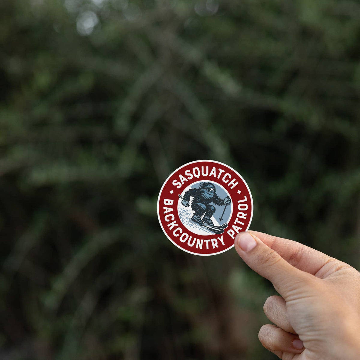 Held at arm's length, the Sasquatch Backcountry Patrol sticker stands sharp against blurred green branches.