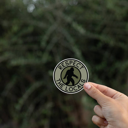 A hand holds the Bigfoot Respect The Locals sticker small in frame against blurred dark evergreen branches.