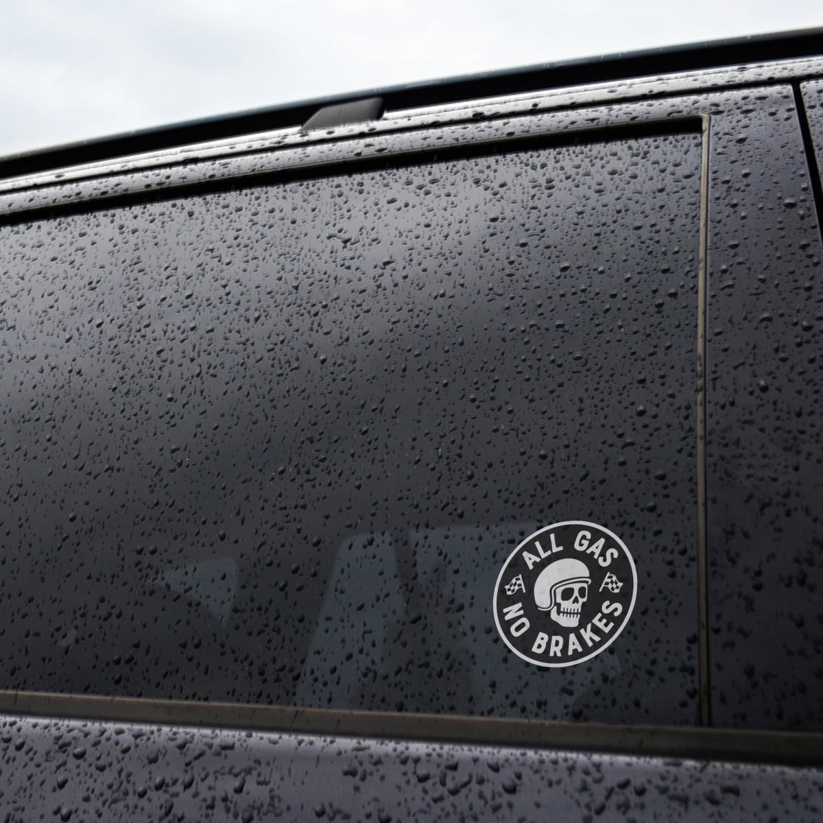 Raindrops bead on a dark car window wearing the All Gas No Brakes sticker near its bottom corner.