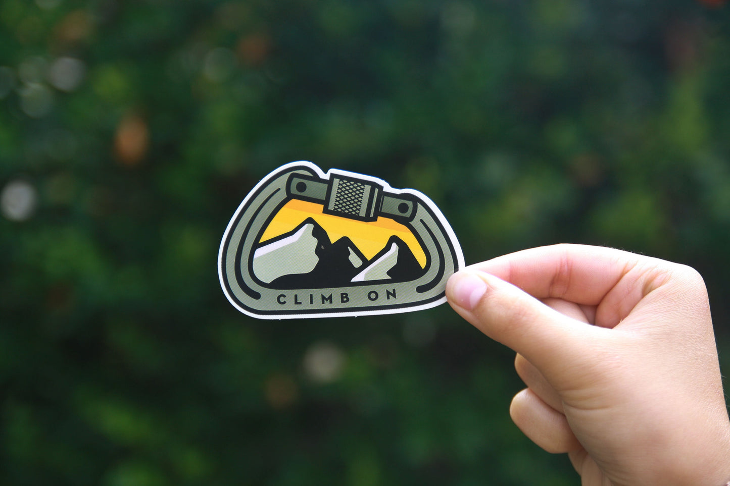 Held up against deep green leaves in soft light, the Climb On Carabiner sticker shows its mountain scene.