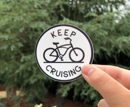 A thumb and finger hold the round Keep Cruising sticker with its beach cruiser bike against a green conifer.