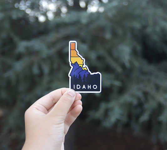 A hand holds the Sawtooth Mountains Idaho sticker against a blurred evergreen, its orange sky above blue peaks.