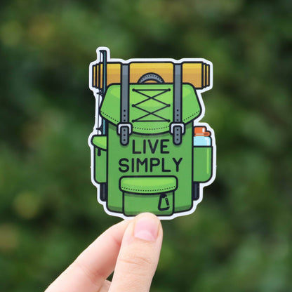 Fingers hold the green Live Simply backpack sticker close to the camera against a blurred wall of leaves.