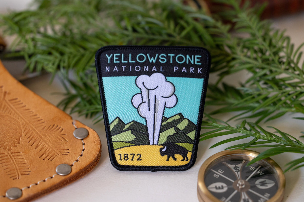 Beside a leather sheath, a brass compass and pine sprigs, the Yellowstone National Park patch rests on white.