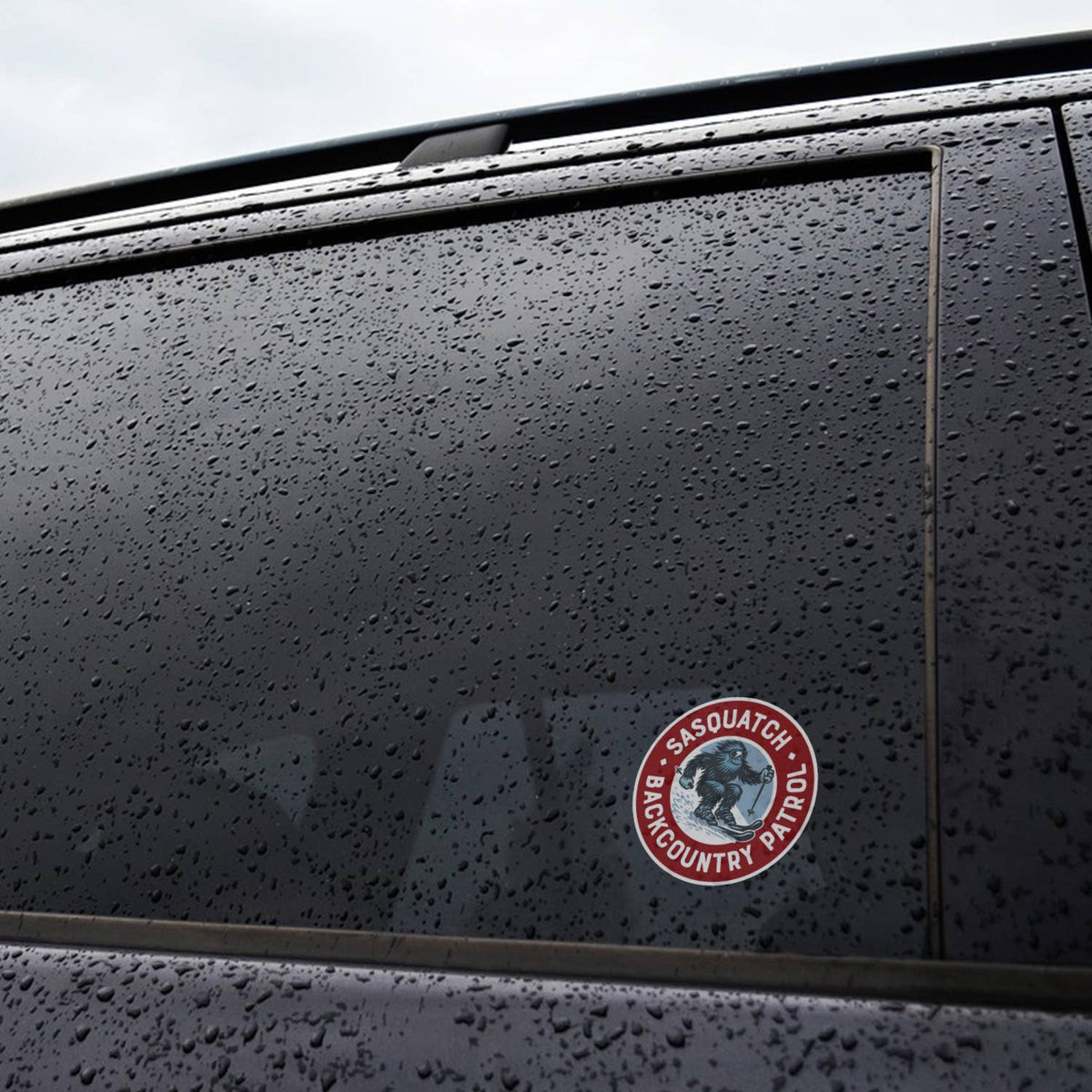 Stuck to a rain beaded car window, the Sasquatch Backcountry Patrol sticker sits low on the dark glass.