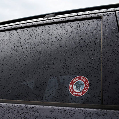 Stuck to a rain beaded car window, the Sasquatch Backcountry Patrol sticker sits low on the dark glass.