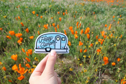 A field of orange poppies sits behind the Happy Camper sticker pinched between finger and thumb.