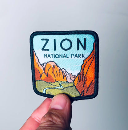 A fingertip lifts the Zion National Park patch toward a soft grey wall, its stitched black edge catching light.