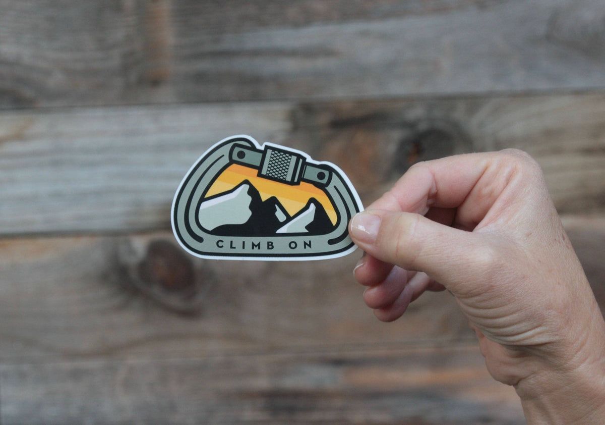 A hand pinches the Climb On Carabiner sticker in front of grey barn boards running horizontal.