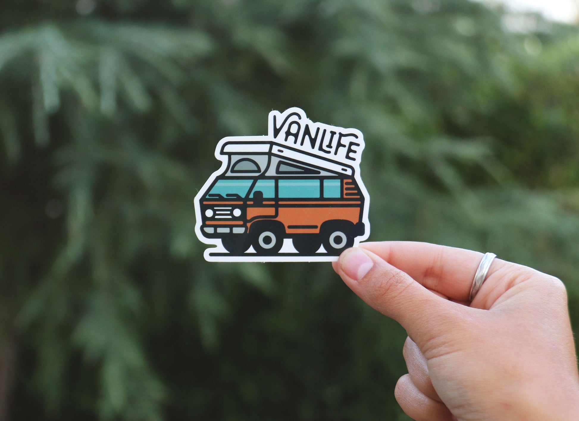 Held further back against a blurred conifer, the Vanlife Pop Top sticker shows its teal windows and pop top roof.