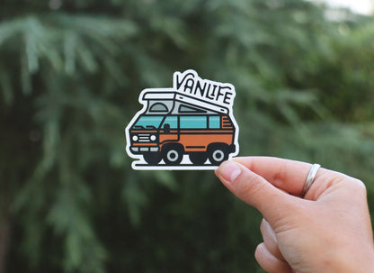 Held further back against a blurred conifer, the Vanlife Pop Top sticker shows its teal windows and pop top roof.