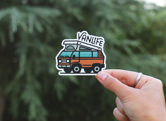 Held further back against a blurred conifer, the Vanlife Pop Top sticker shows its teal windows and pop top roof.