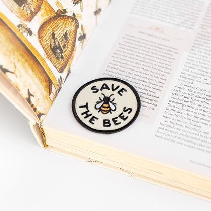 Resting on a bee illustrated book page, the round Save The Bees patch sits near the spine's cream edge.