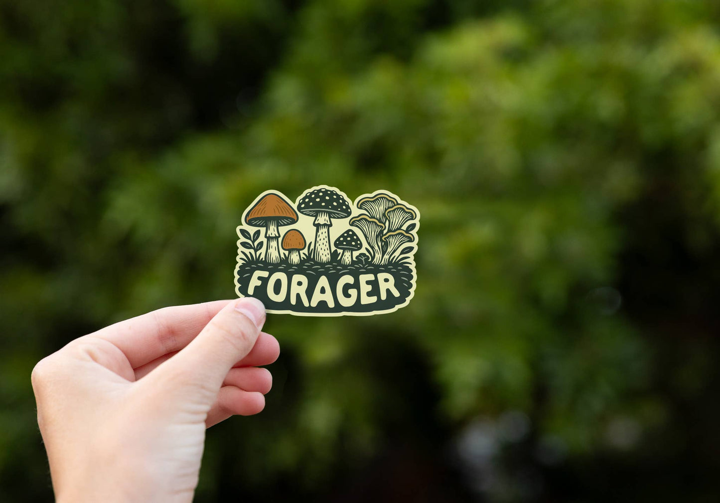 A hand holds the small Forager Mushrooms sticker against a blurred wall of dark green foliage.