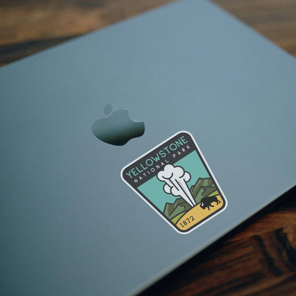 Stuck near the apple on a blue laptop lid, the Yellowstone National Park sticker sits above dark wood.