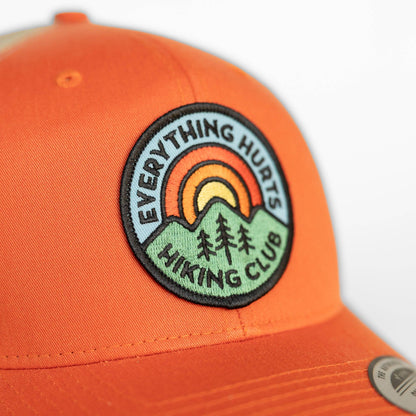Sunrise arcs in orange and yellow on the patch stitched to the orange Hiking Club trucker hat crown.