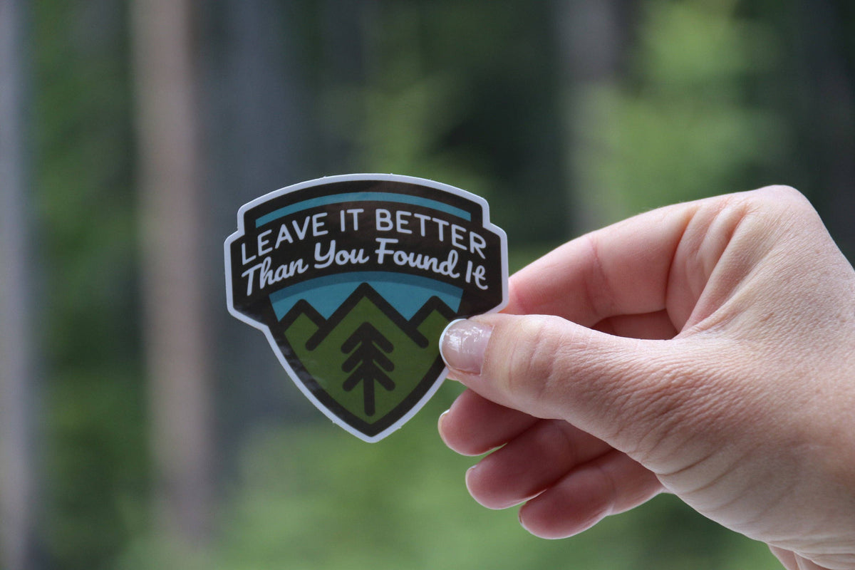 Fingers grip the Leave It Better Than You Found It sticker with blurred green forest behind.